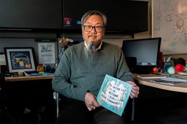 David Lo sitting at desk holding comic book. Picture taken by Kitra Cahana for Undark